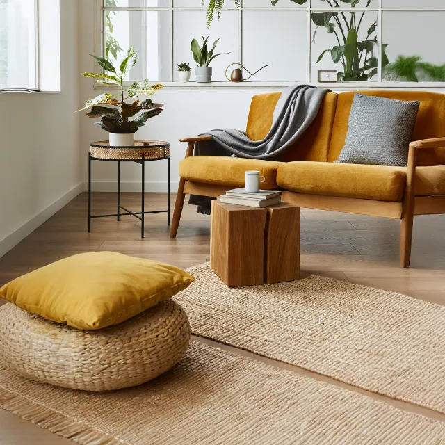A living room with a mustard sofa, a cane side table, an oak stool and a woven pouffe